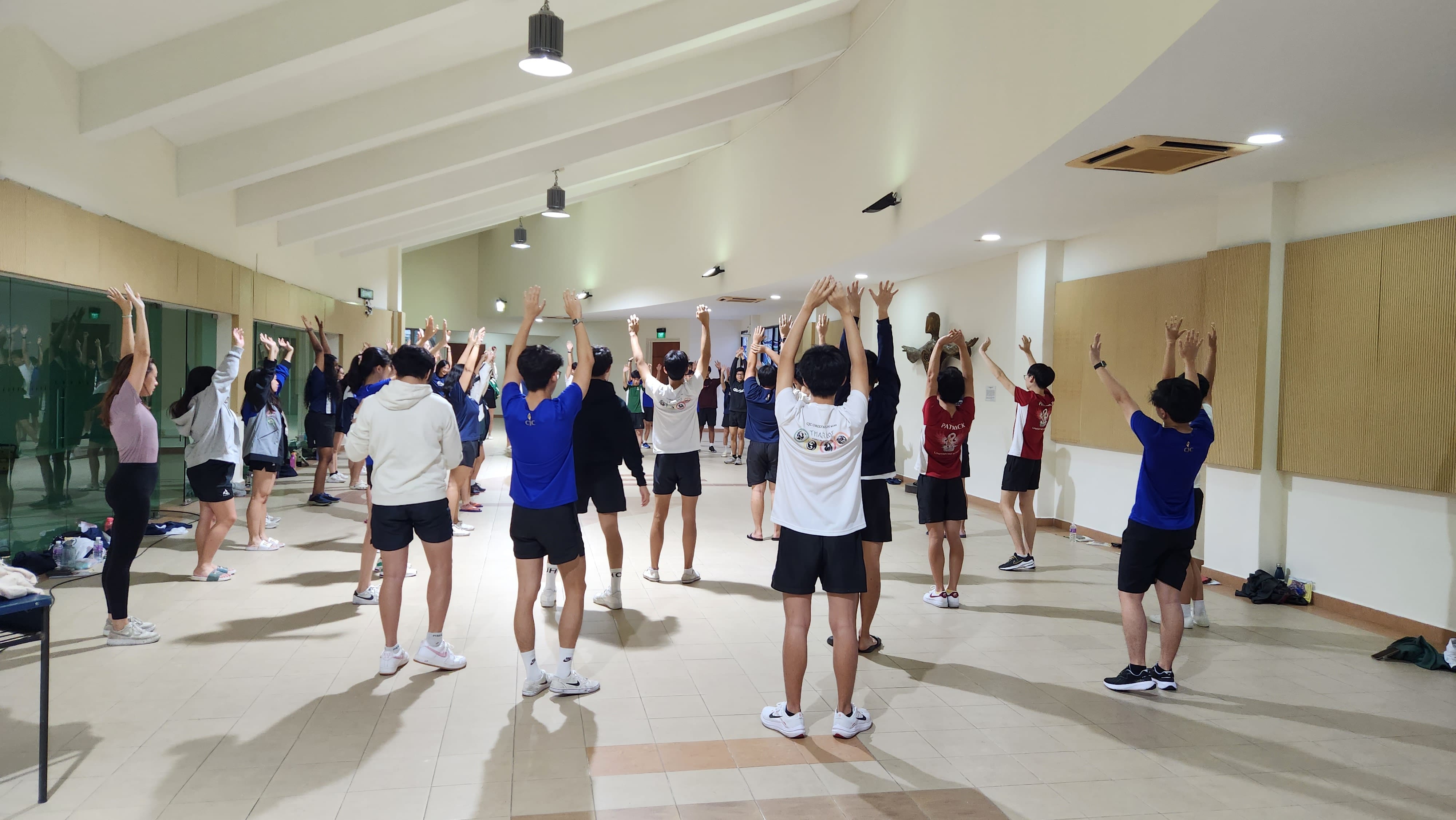 A large group of students in sportswear stretch their arms overhead in a brightly lit indoor hall.